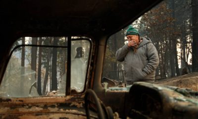 Photo of a wildfire victim wiping his eyes as he walks by a burned car