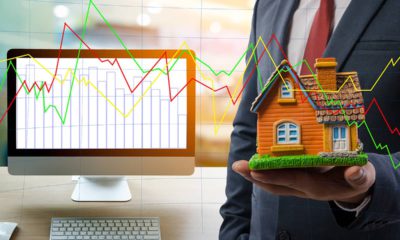 Photo of man holding a house next to a computer with stock charts