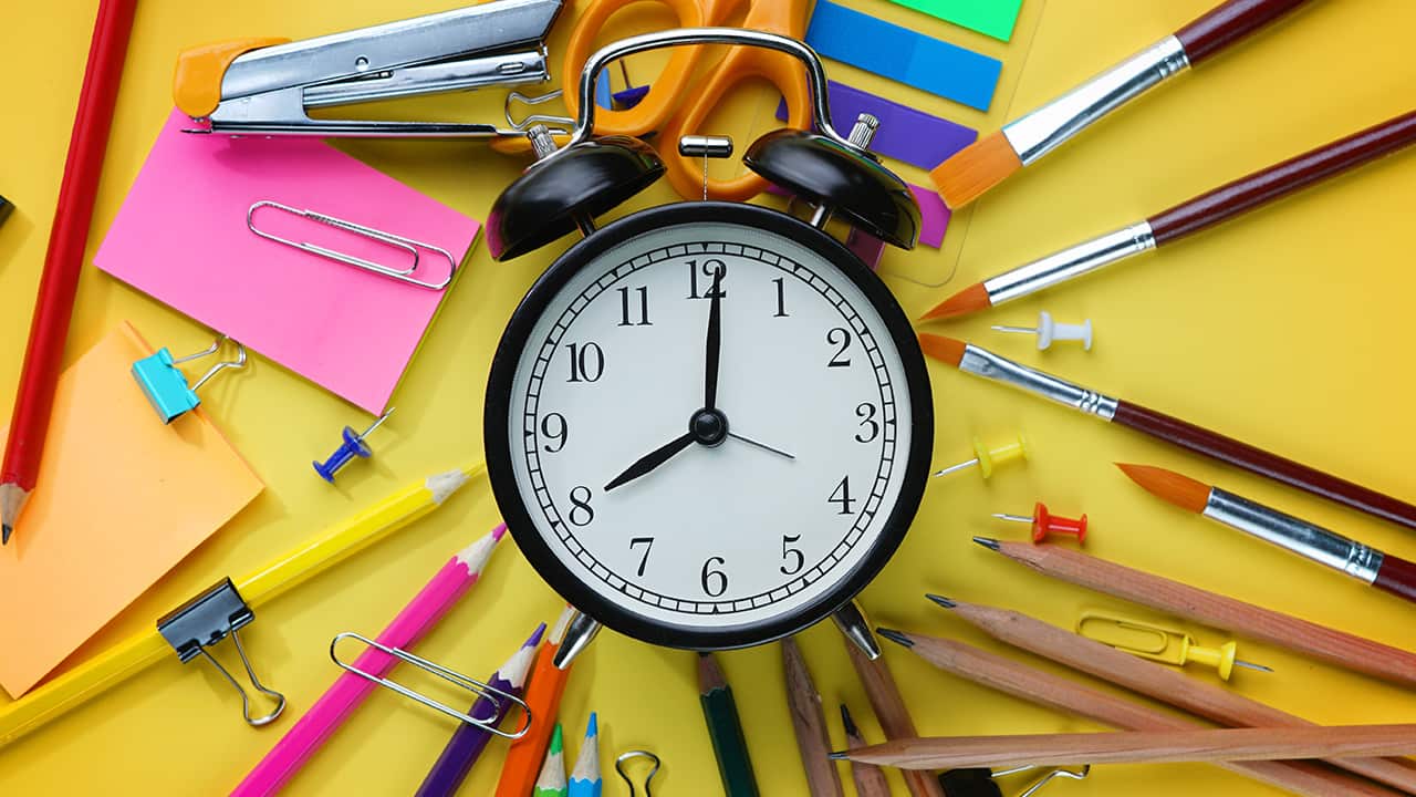 Photo of alarm clock against a backdrop of pens, rulers and paper signifying school start timesr